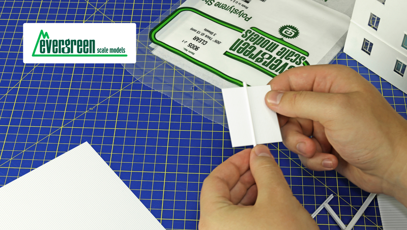 Hands assembling a white styrene model building on a cutting mat, beside an Evergreen product pack